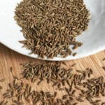 Cumin seeds in a white bowl overflowing onto a wooden board.