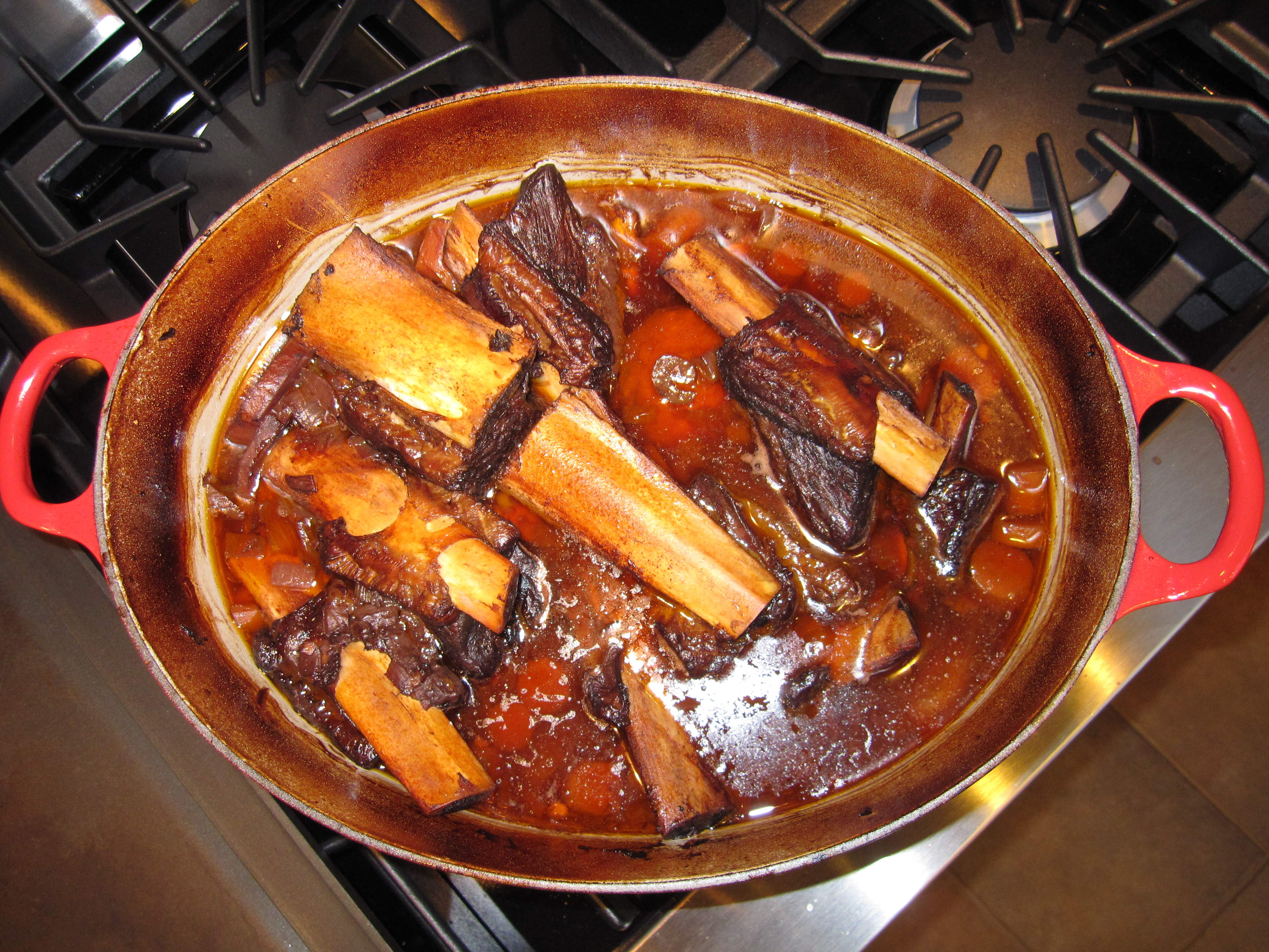 Dutch oven full of vegetables, short ribs and sauce out of oven, sitting on stove top.