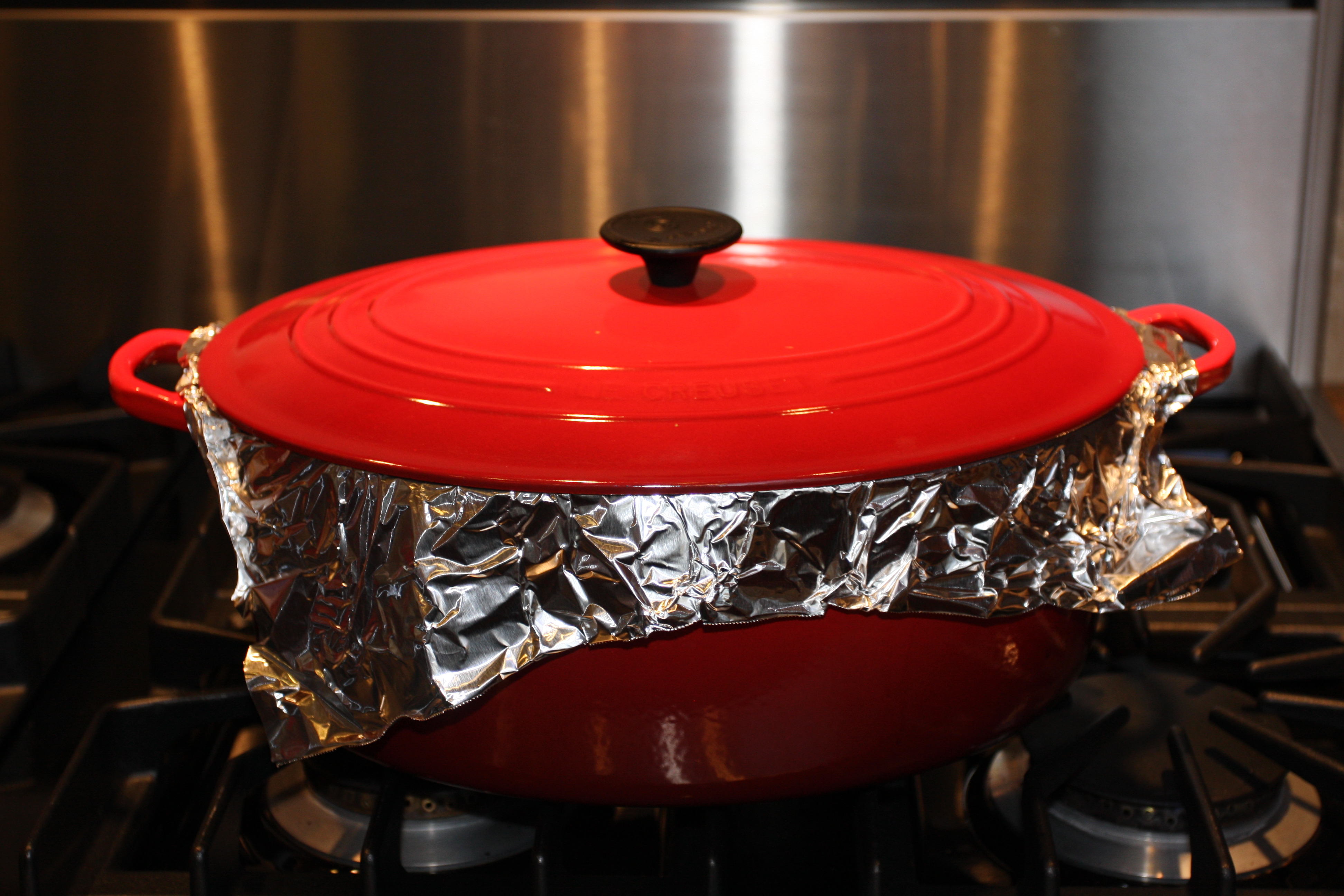 Red Dutch oven with foil over top and lid placed on top.