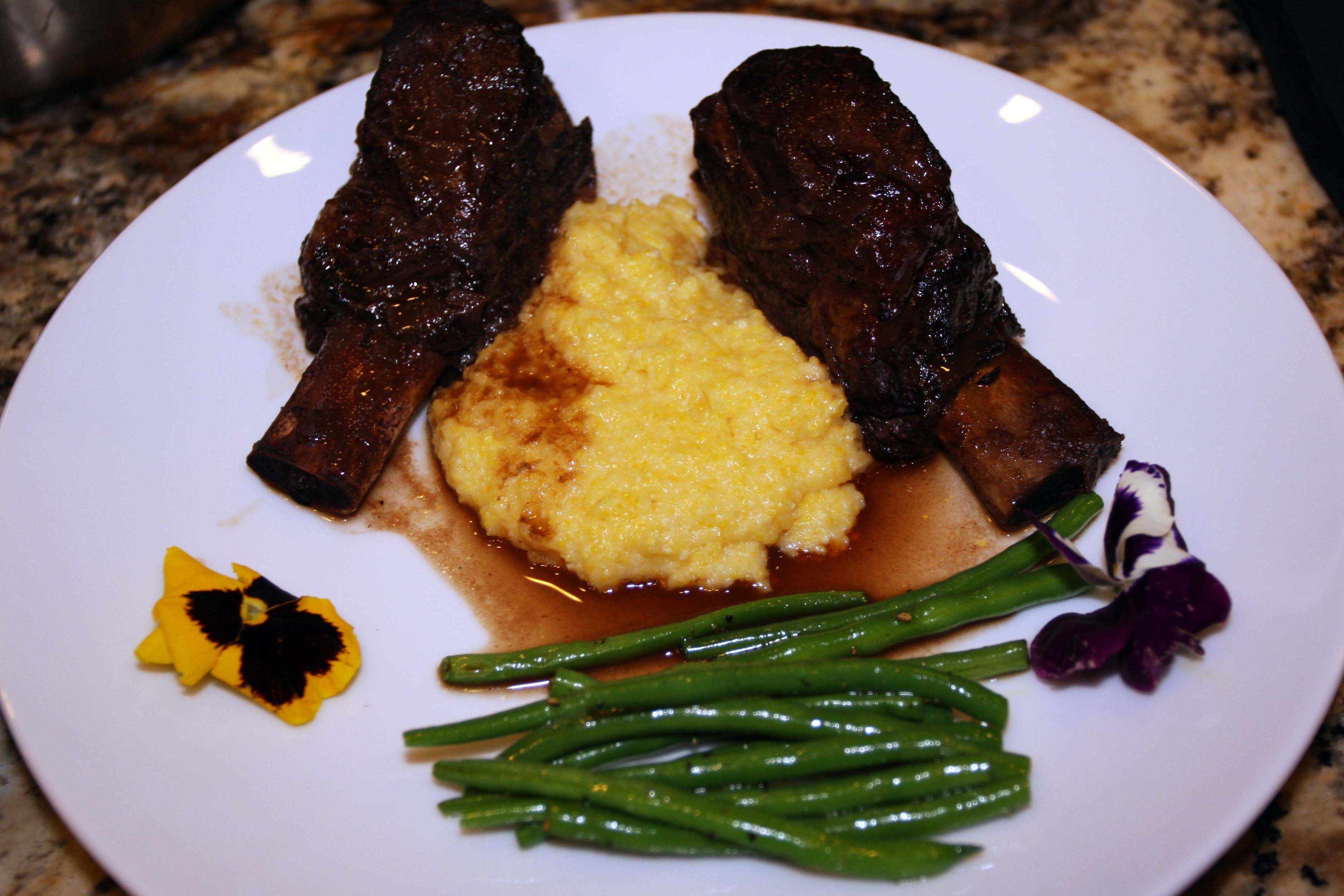White plate of short ribs, mashed potatoes, and haricot verts garnished with edible flowers.