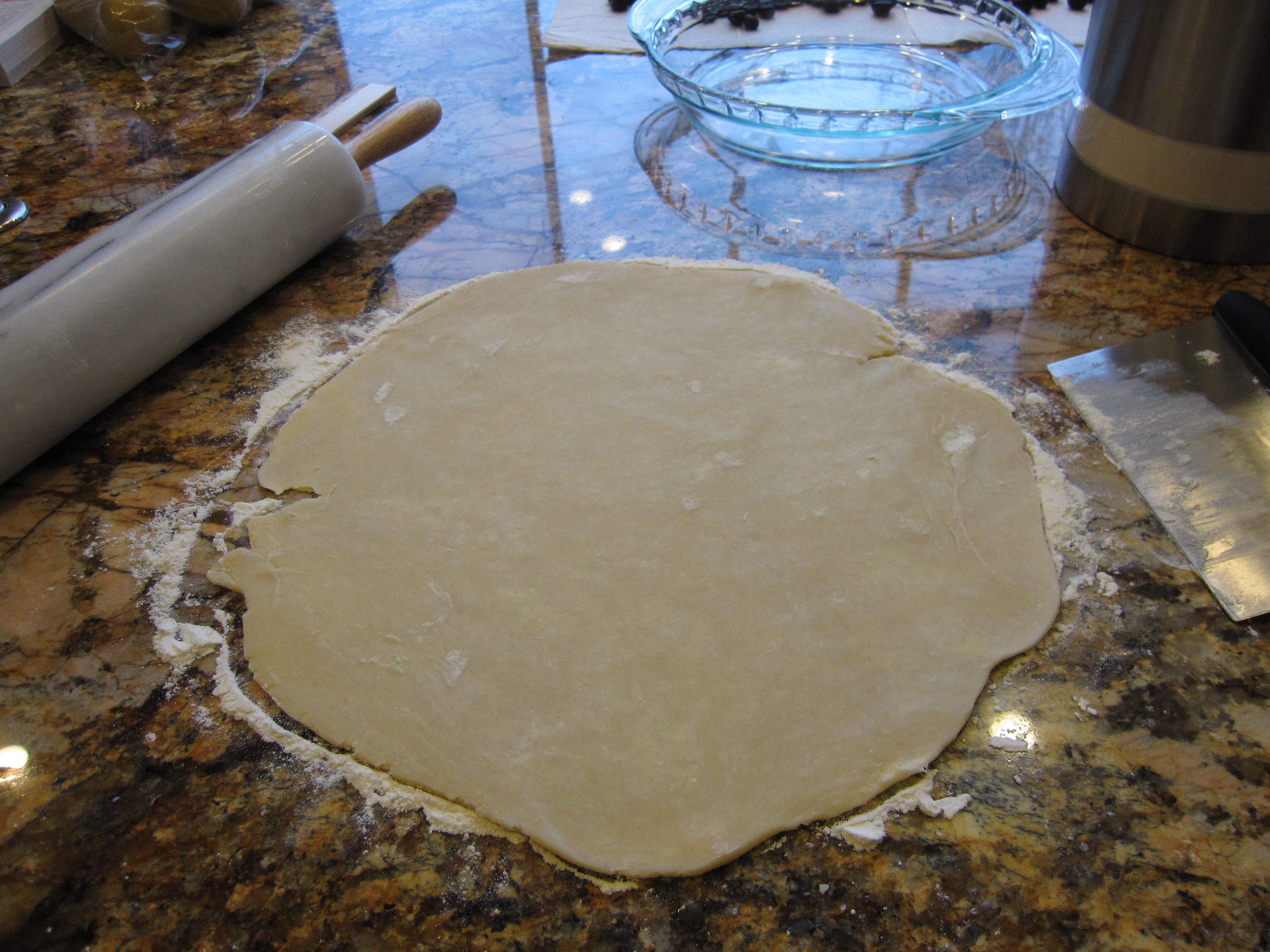 Pie dough rolled out into a circle on a counter top with bench scraper and rolling pin on hand. 