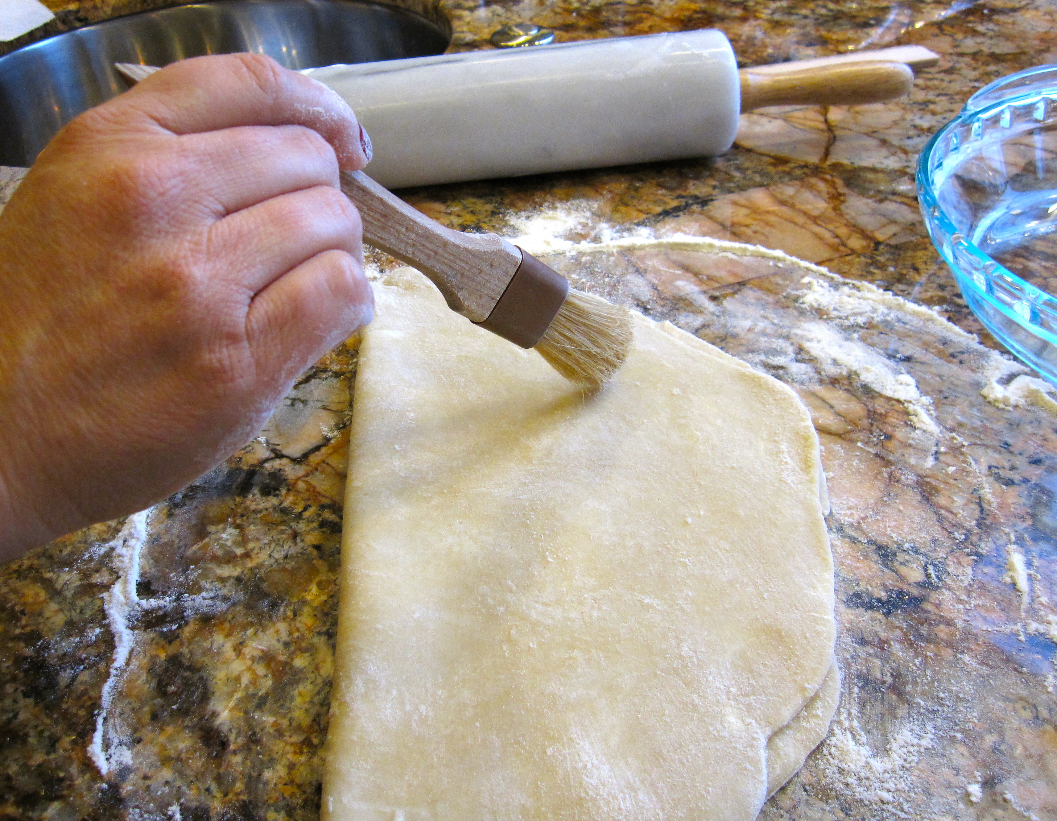 Using a brush, removing excess flour from folded in half circle of pie dough on counter.