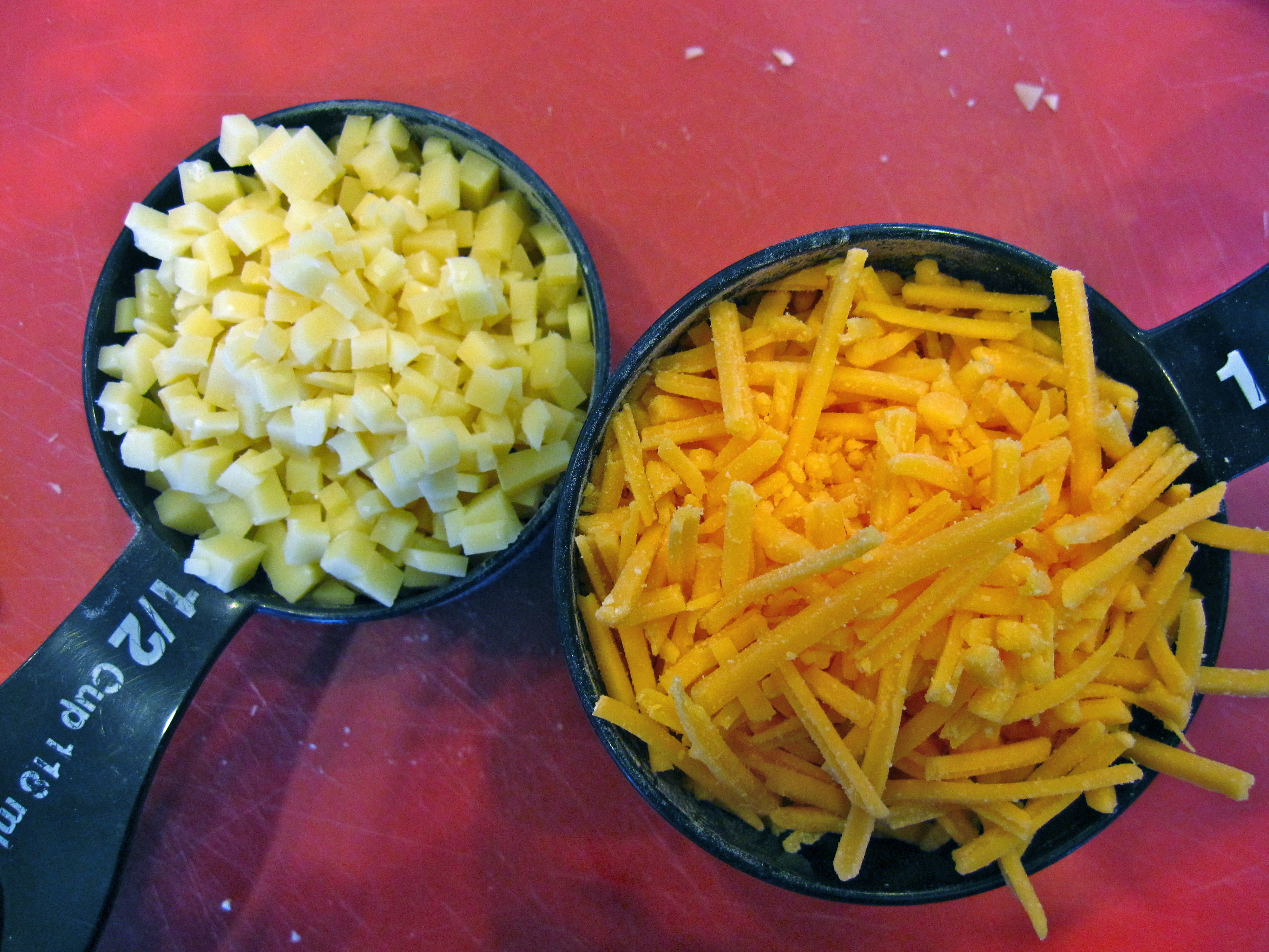 2 black measuring cups, one filled with cubed grana padano cheese, the other with shredded cheddar cheese. 