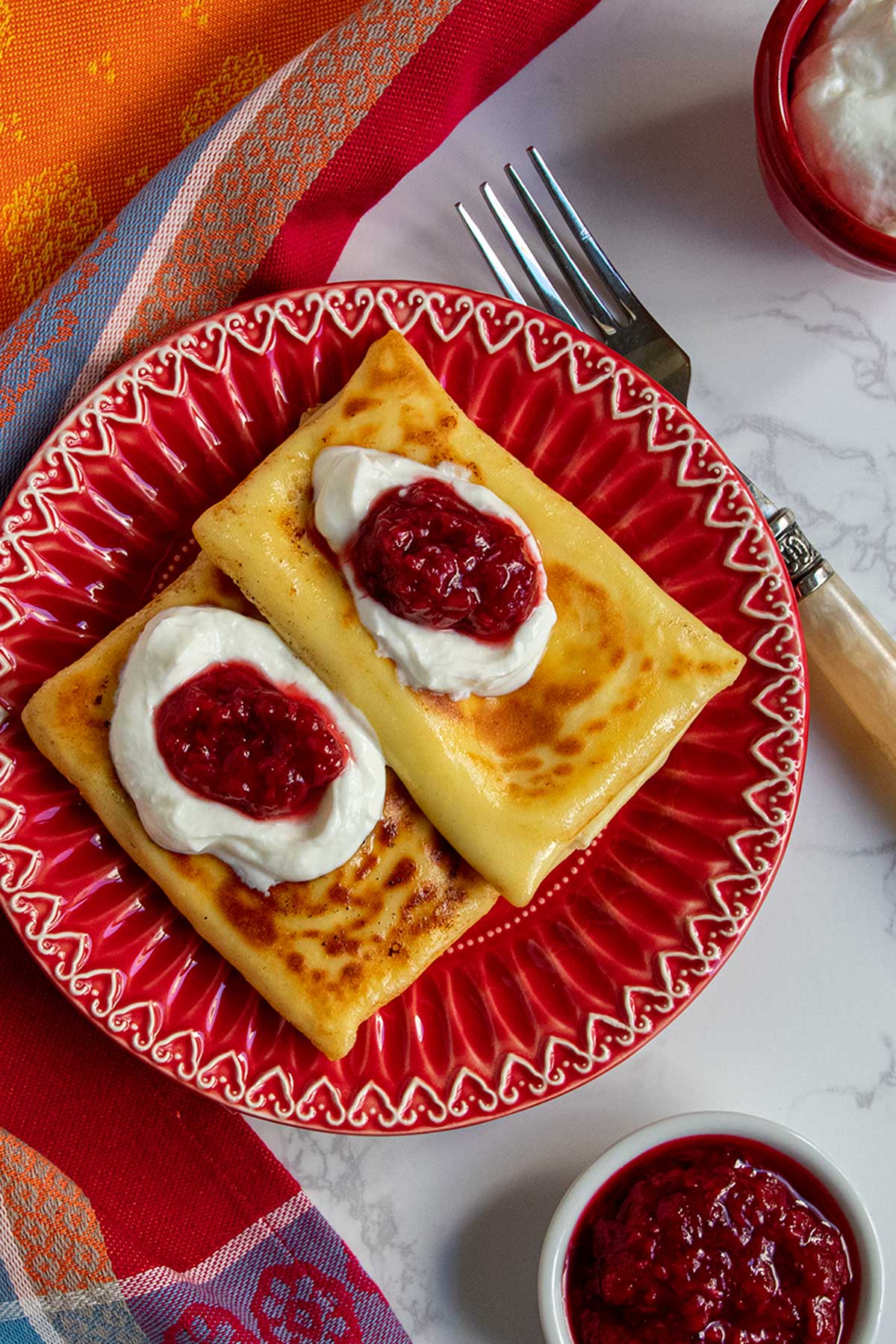 2 blintzes on red plate topped with sour cream and jam.