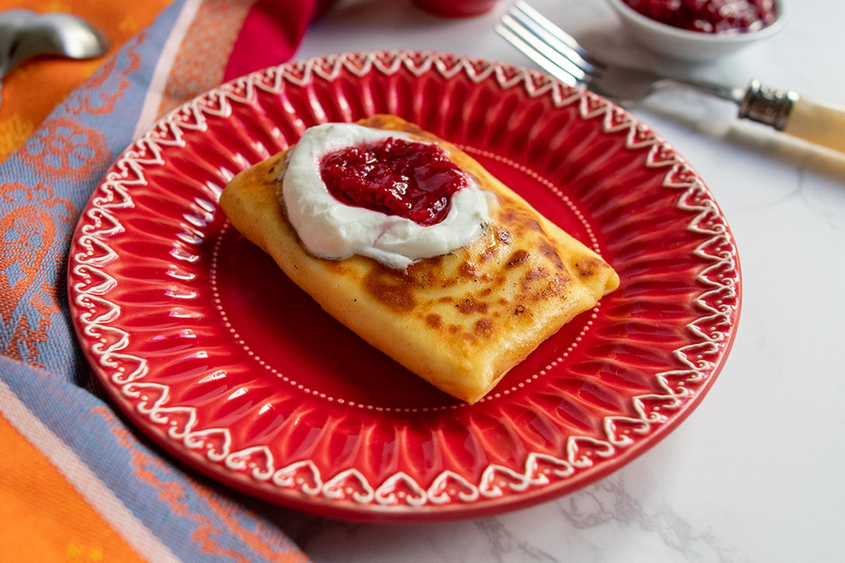 One blintz on red plate topped with jam and sour cream.