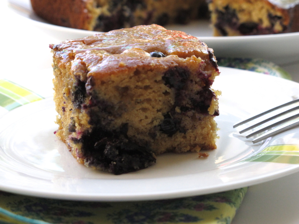 A serving of Melissa Clark's maple blueberry teacake or pancake? Square piece showing the blueberries and moist cake on a white plate with a fork.