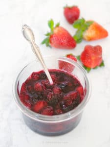 fruit compote in a glass bowl with silver spoon and fresh strawberries on the side
