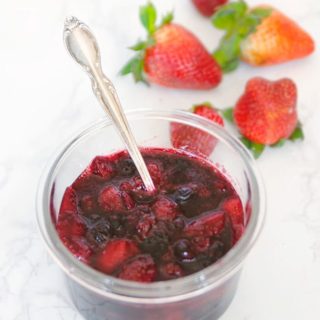 fruit compote in a glass bowl with silver spoon and fresh strawberries on the side