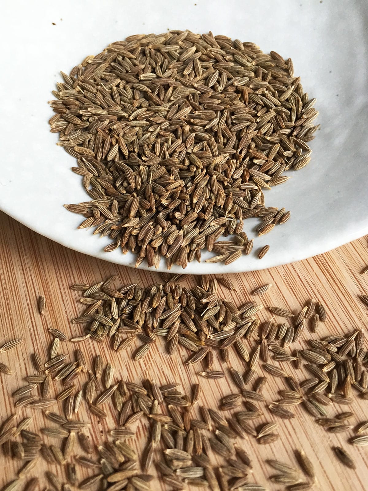 Cumin seeds in a white bowl overflowing onto a wooden board.