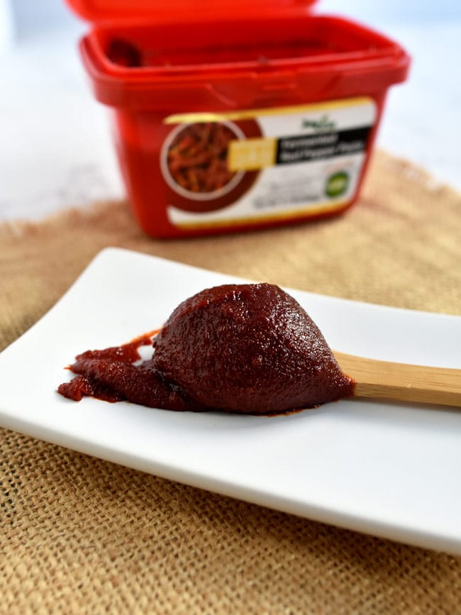 Deep red Gochujang spice paste on a wooden spoon with red container in the background.