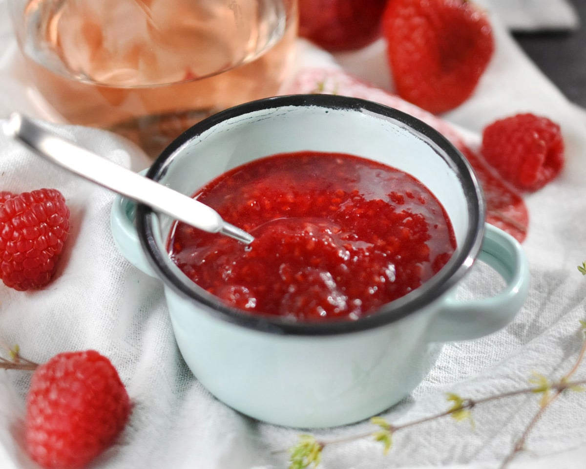 Gregory's jam in small bowl with spoon surrounded by fresh raspberries.