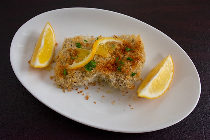 A serving of breaded chicken on white plate and dark background garnished with fresh lemon wedges and a twist of lemon on top.
