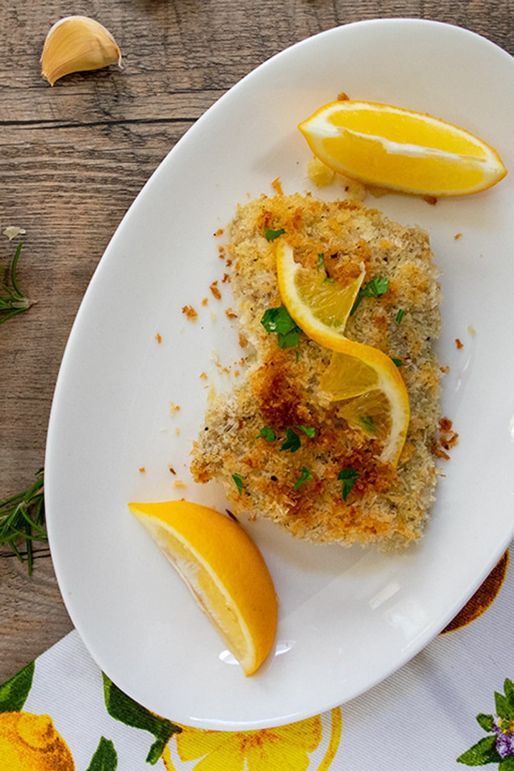 Breaded lemon chicken on a white plate with a head of garlic, a cut lemon and  sprig of fresh rosemary next to the serving. 