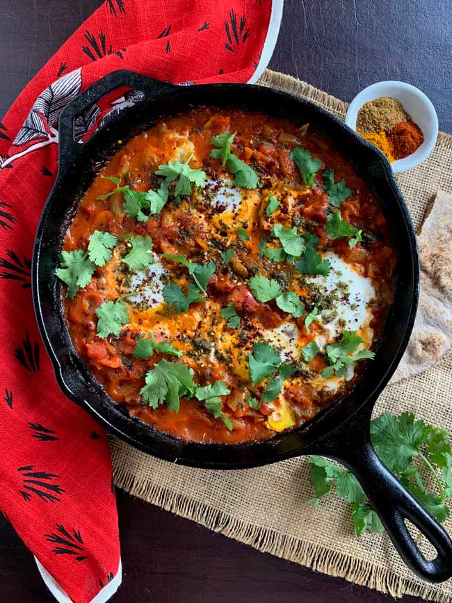 Full pan view of shakshuka with pita bread and cilantro and spices.