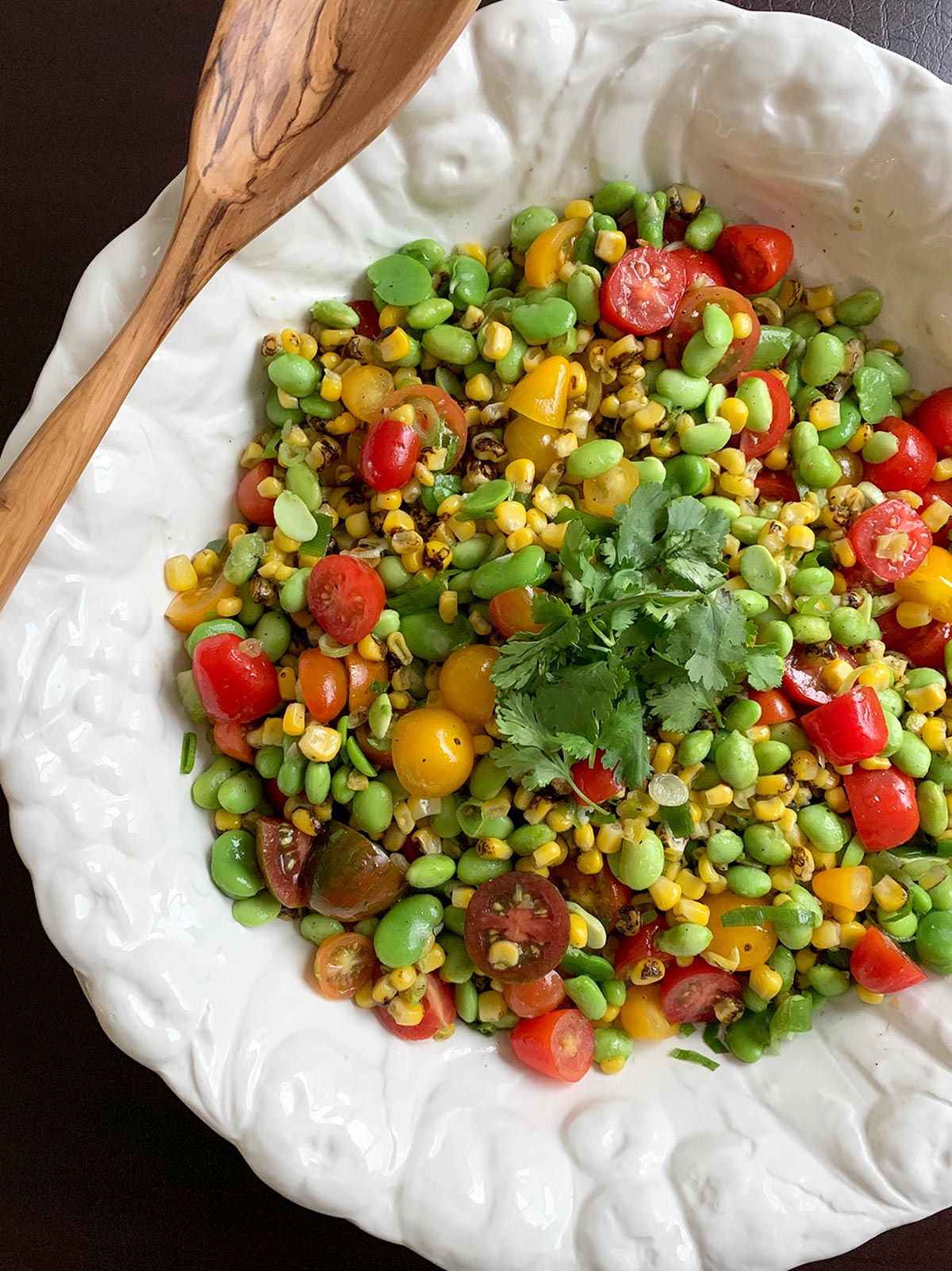 White bowl with corn salad in it and a large wooden spoon on top.