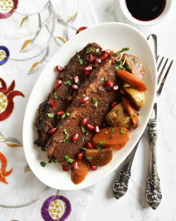 Pomegranate molasses brisket on a white plate with carrots and potatoes on a pomegranate design napkin.