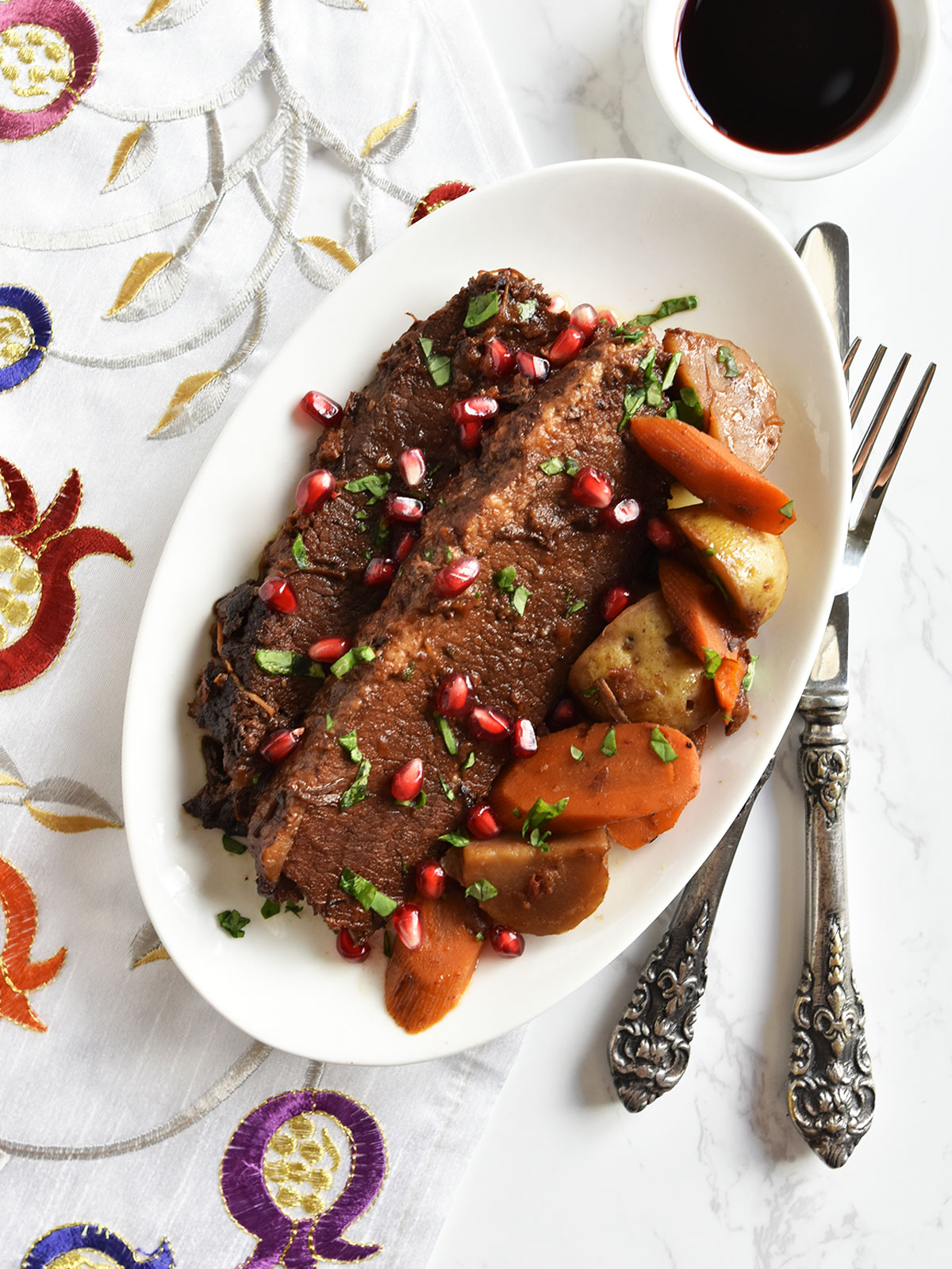 Pressure cooker brisket with pomegranate molasses on a white plate with carrots and potatoes on a pomegranate design napkin.
