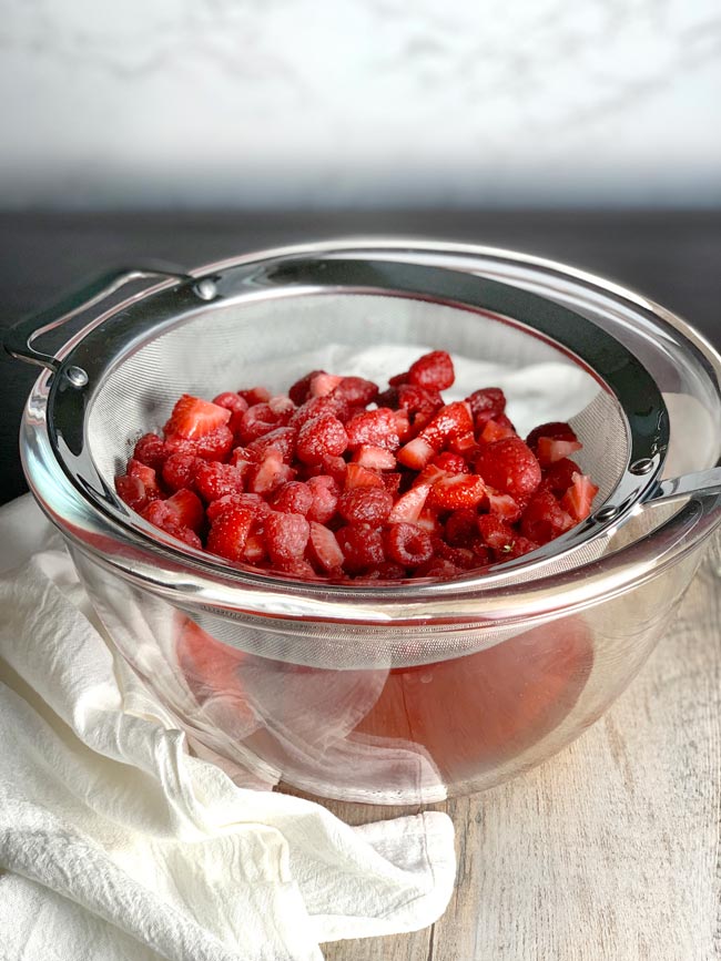 Rosé jam ingredients in a metal strainer over a bowl,  straining liquid out. Cheesecloth is next to the bowl.