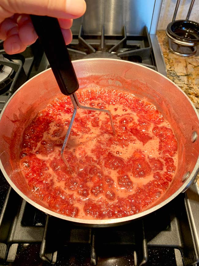 Mashing strawberry raspberry jam in pot on stovetop while boiling. 