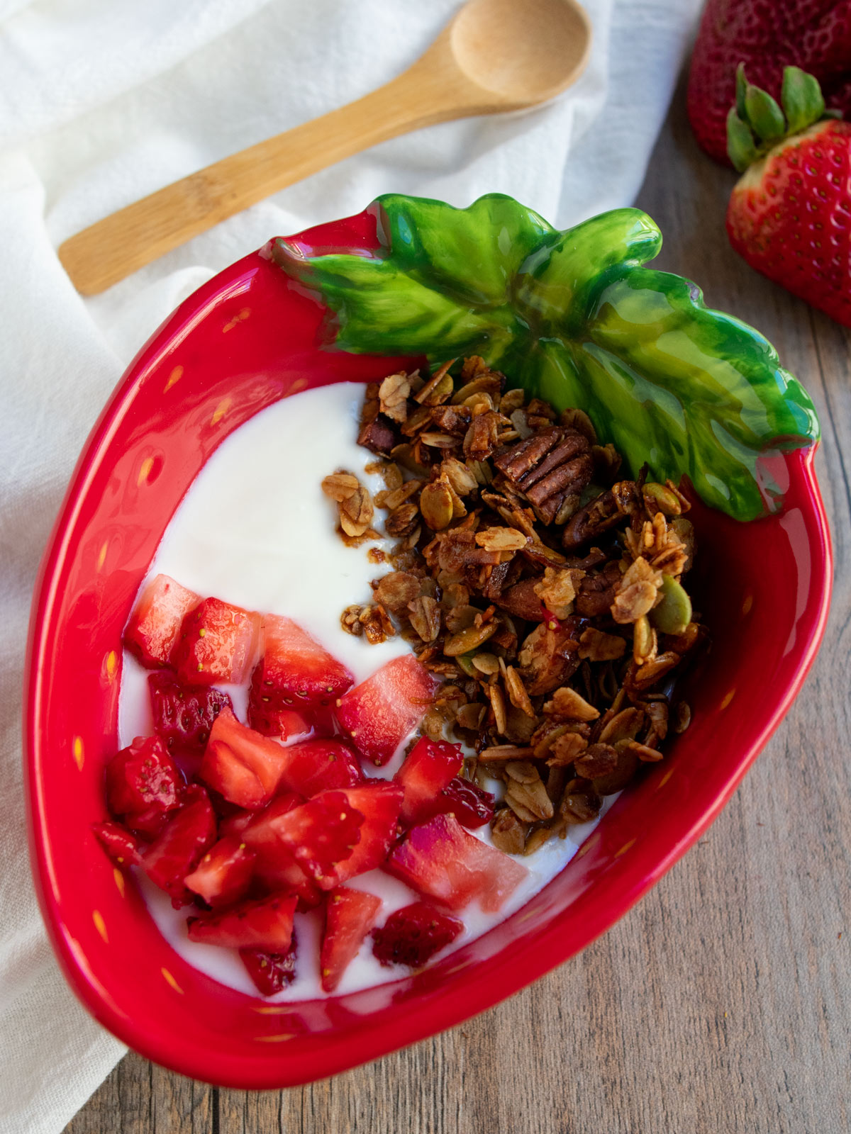 Olive oil granola in a strawberry red bowl on top of yogurt with strawberries on top as well.