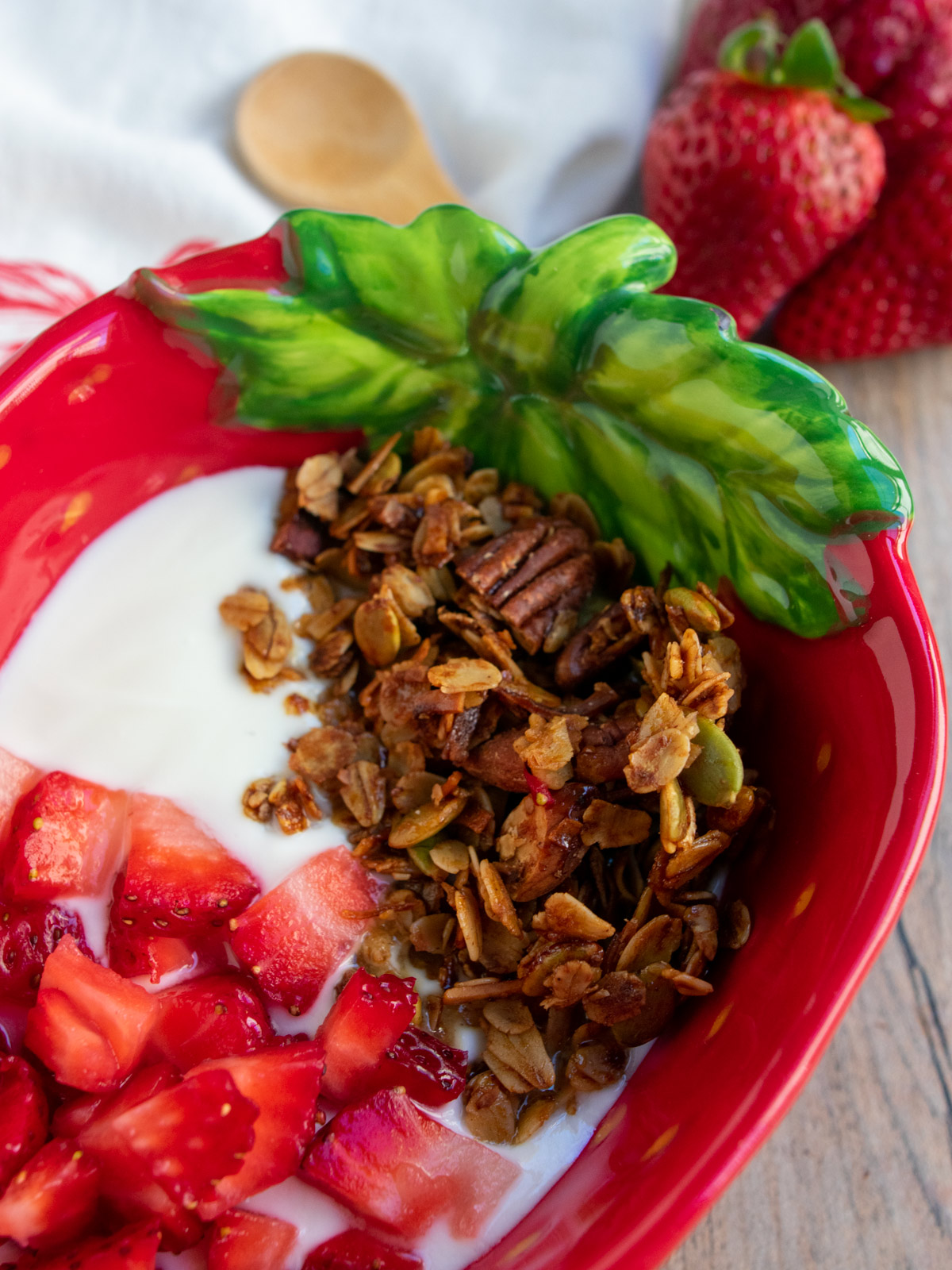 Close up of olive oil granola in the strawberry bowl with fresh strawberries and a spoon behind it.