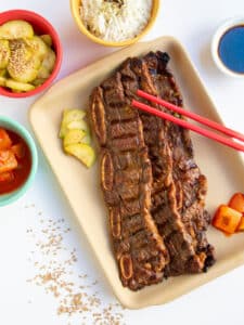 Kalbi ribs on a tan plate with red chopsticks and various banchan on the side.