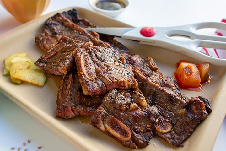 Kalbi ribs cut into small pieces using kitchen shears sitting on tan platter with cucumber and kimchi.