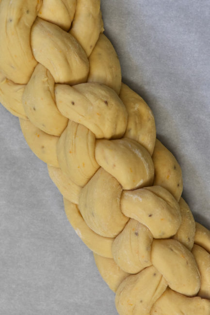 Unbaked challah braided as 6 strands laying on white parchment paper.