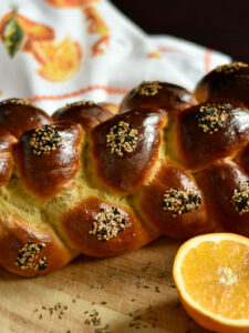 Side view of a baked loaf of challah with seeds on it and an orange in the foreground.