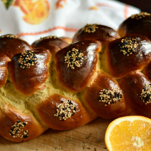 Side view of a baked loaf of challah with seeds on it and an orange in the foreground.
