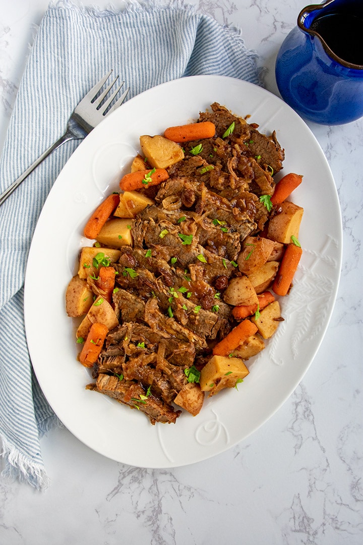 Large white platter of slow cooker brisket, potatoes, carrots. Served along with striped napkin, fork and blue pitcher of gravy.