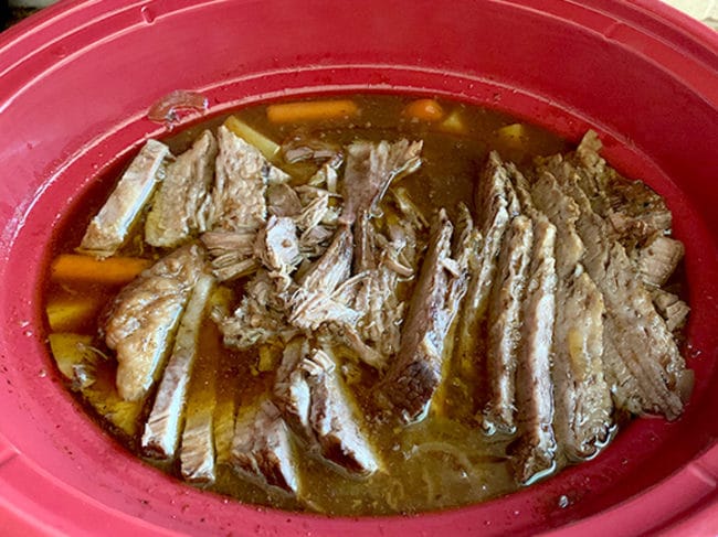 Finished and sliced brisket and vegetables still in red slow cooker.