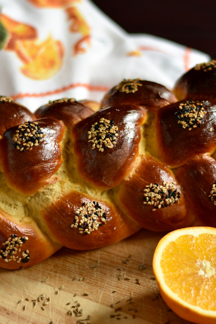 Close up of braided challah with a sliced orange in the foreground and a white napkin with oranges on it in the background.