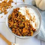 Pumpkin pie granola in a bowl with yogurt and white pumpkin on the right and a spoon on the left with yogurt and granola on it.