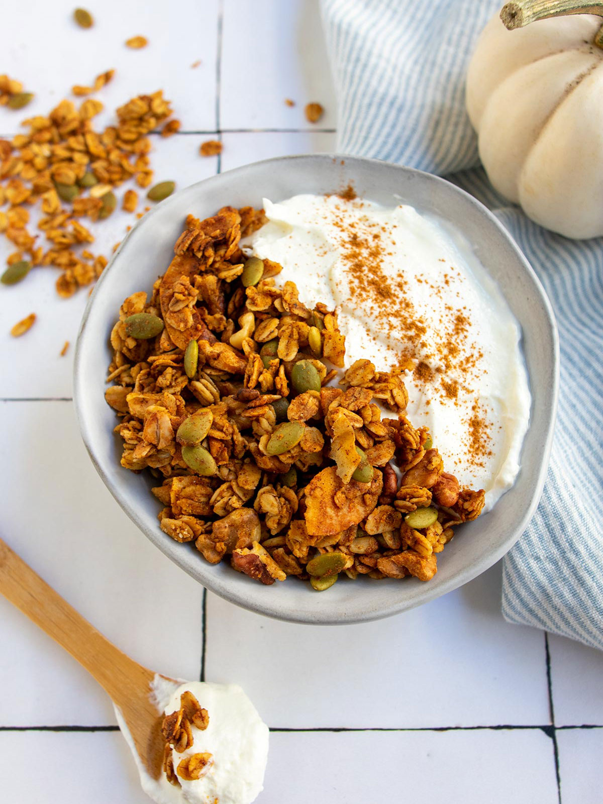 Pumpkin pie granola in a bowl with yogurt and white pumpkin on the right and a spoon on the left with yogurt and granola on it.