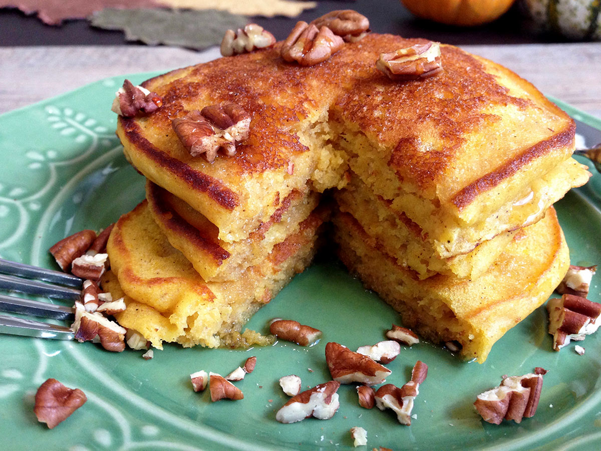 Stack of pumpkin pancakes on green plate with pecans and maple syrup.