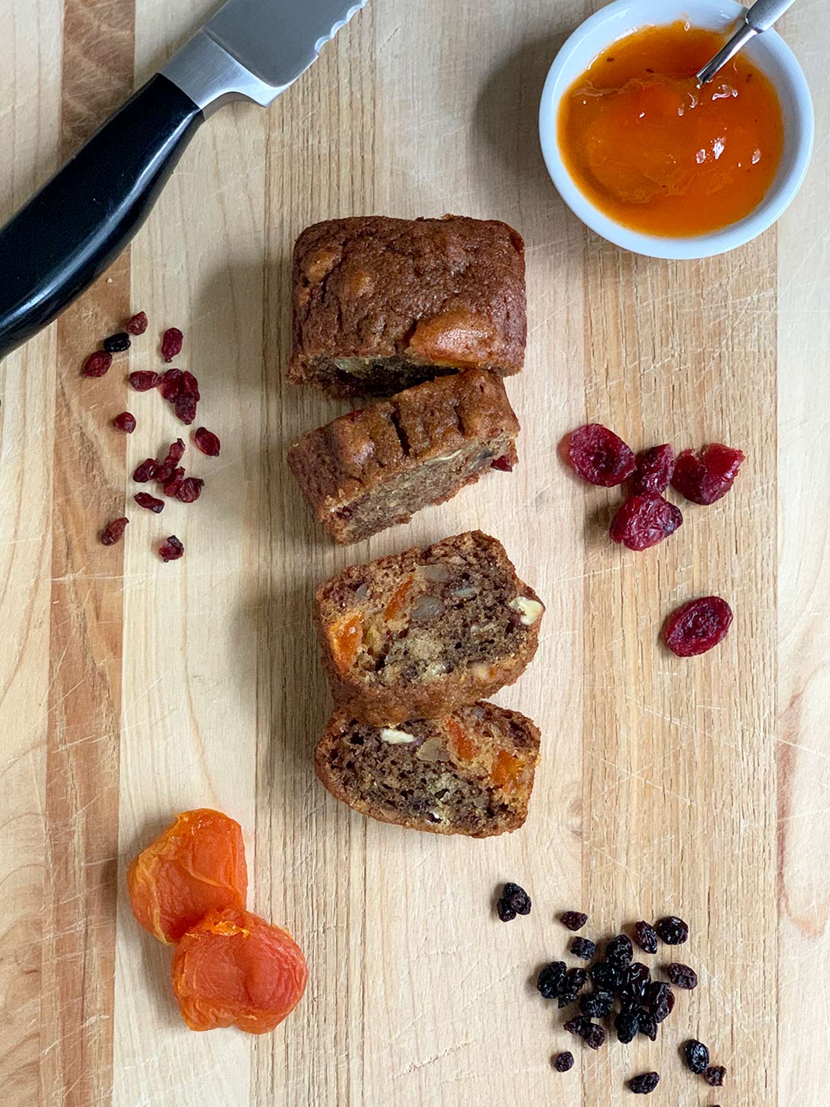 Mini persimmon loaf sliced on cutting board along side dried fruit, persimmon pulp, and a knife.