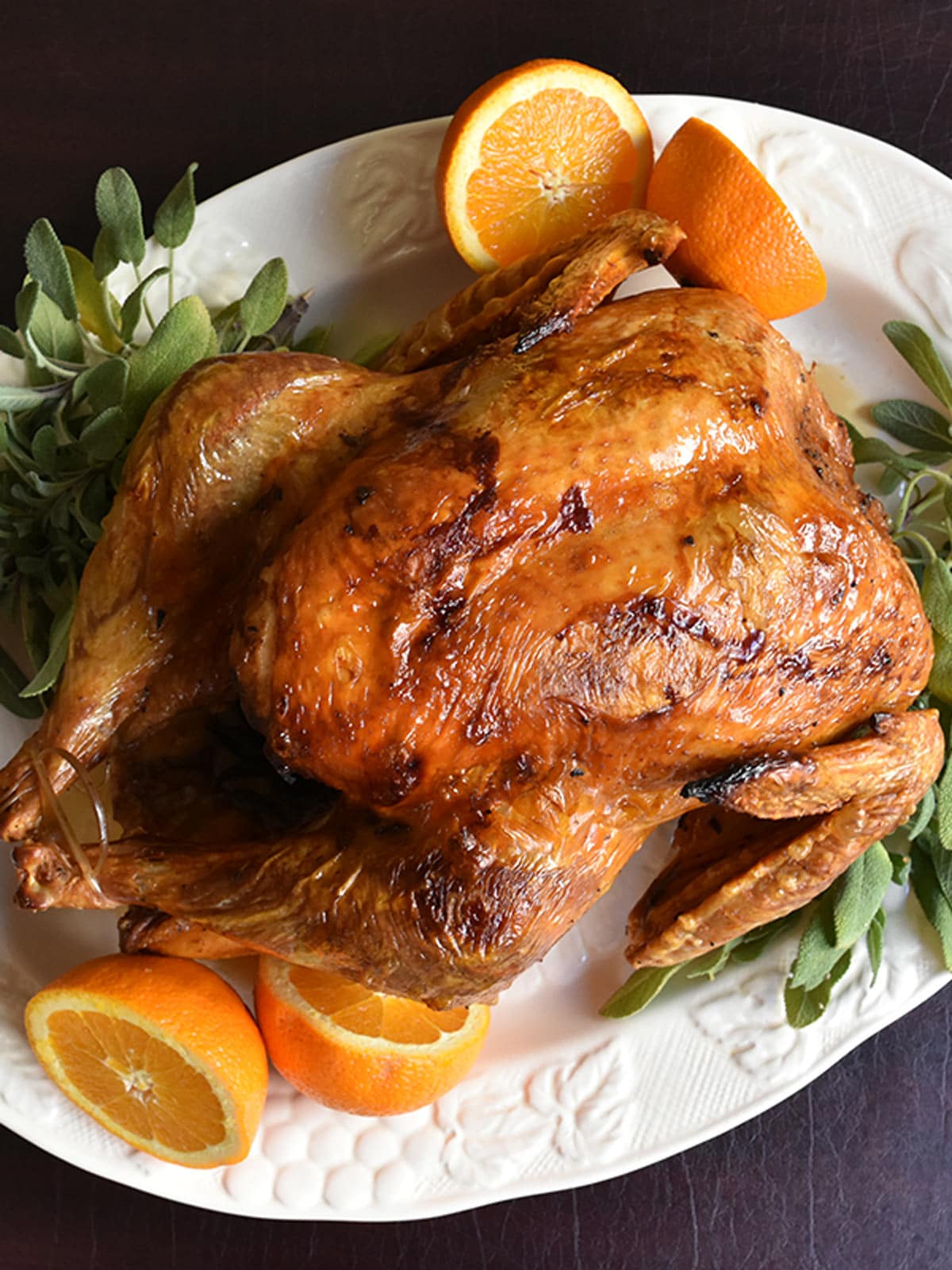 Top down view of golden brown roasted turkey on white platter with sage and orange garnish.
