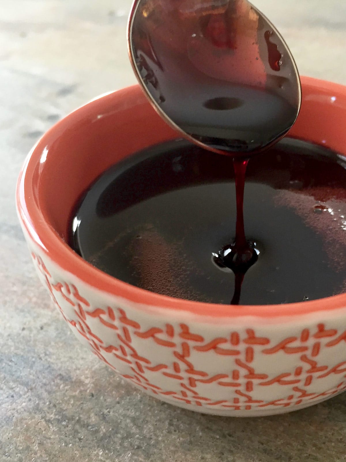Dark syrupy pomegranate molasses dripping off of a spoon into an orange bowl.
