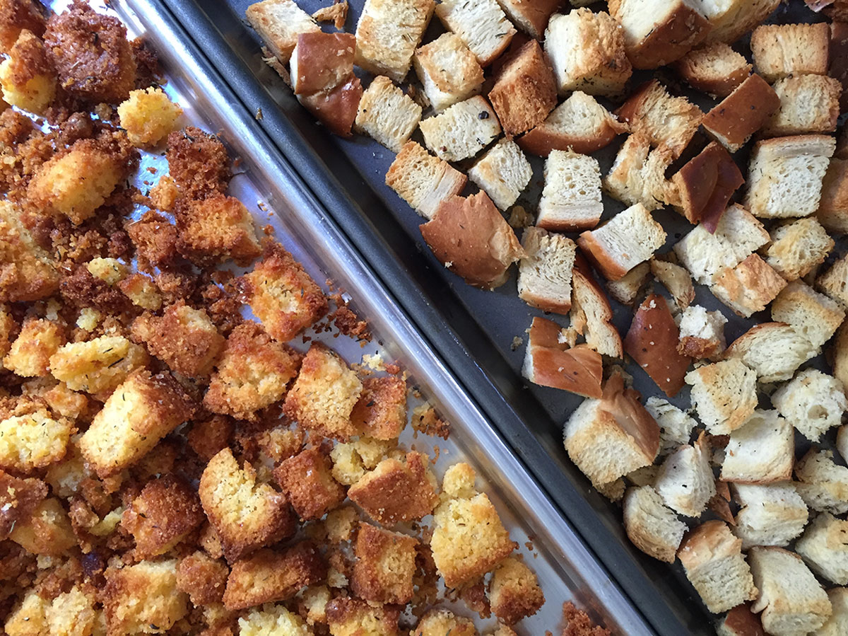 Cornbread and challah stuffing bread cubes on sheet trays at an angle.