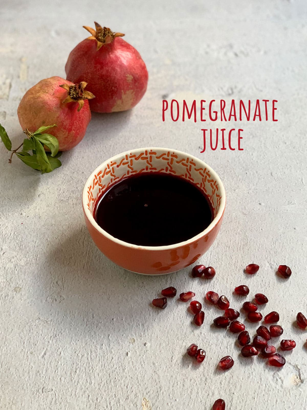 A bowl of dark red pomegranate juice next to a scattering of seeds and 2 whole pomegranates.