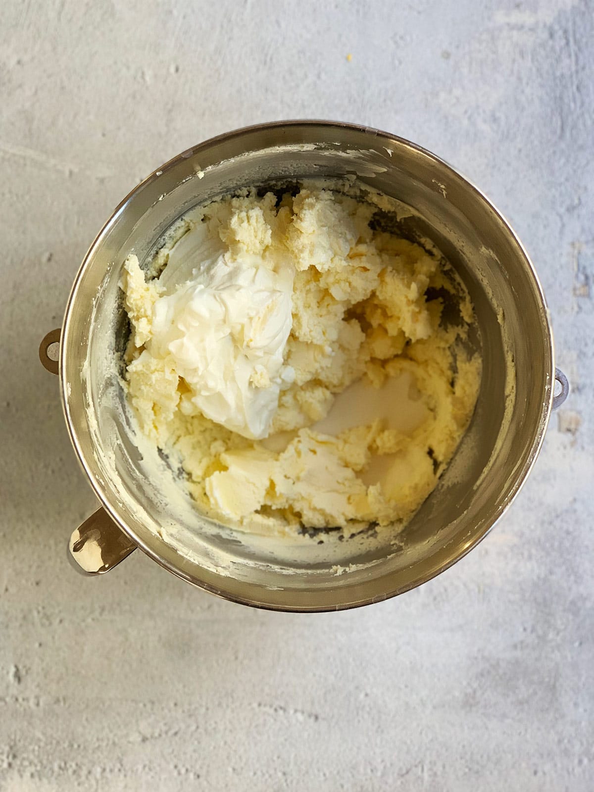 Sugar and sour cream added to whipped butter and cream cheese mixture in a metal mixing bowl.