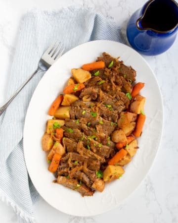 Straight down view of large white platter with slow cooker sliced brisket and potatoes and carrots on it plus a serving fork on the side and a pitcher of gravy.