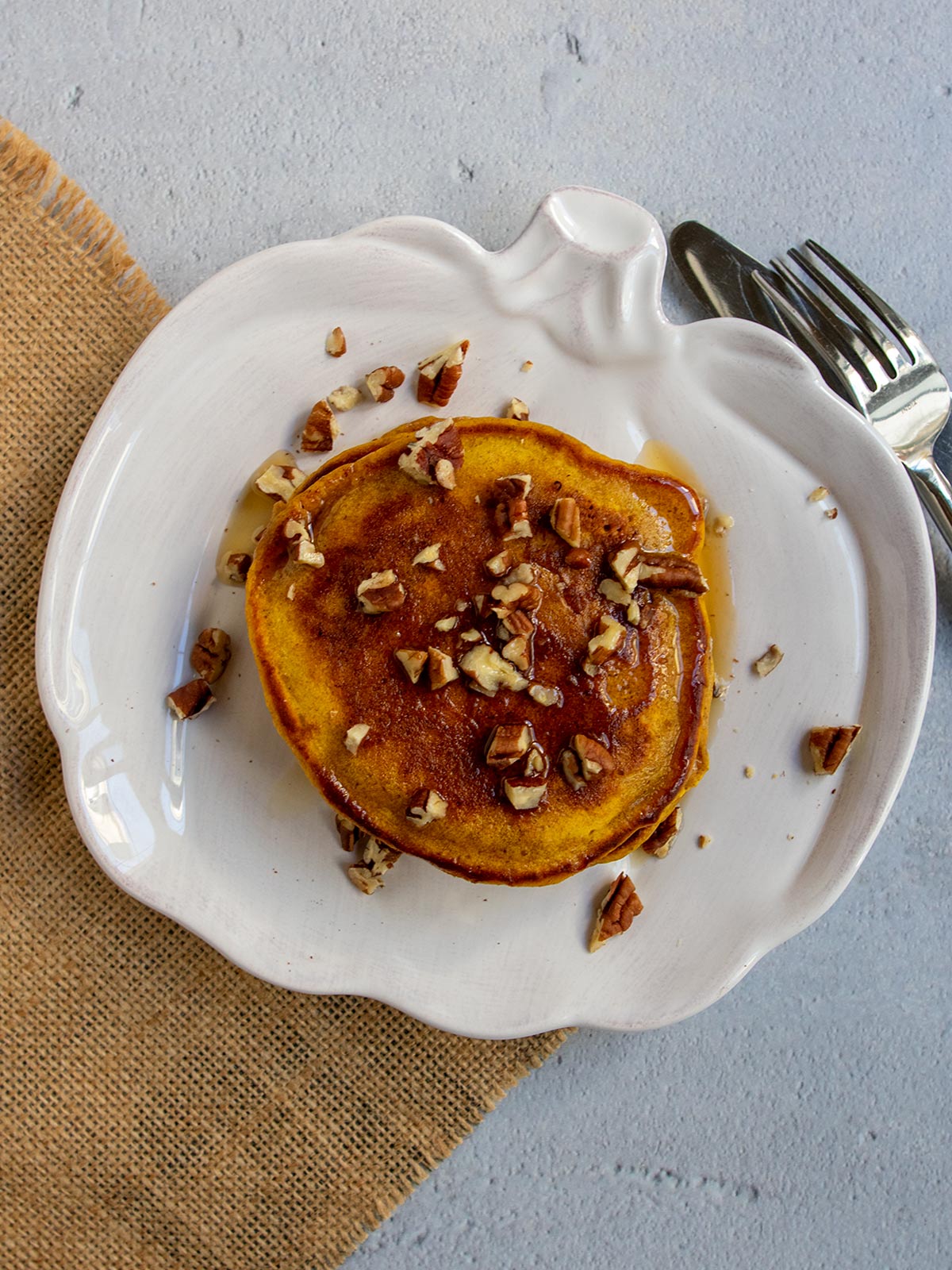 Stack of pumpkin spice pancakes on pumpkin-shaped plate topped with pecans and maple syrup.