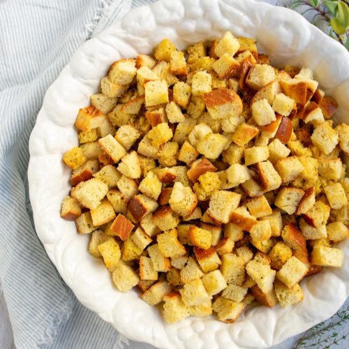 Large white bowl with bread cubes in it and a striped napkin and fresh herbs on the side.