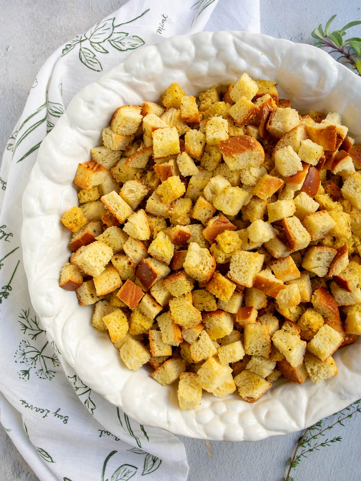 Cornbread and challah stuffing cubes in a white bowl with a herb decorated towel and fresh herbs on the side.