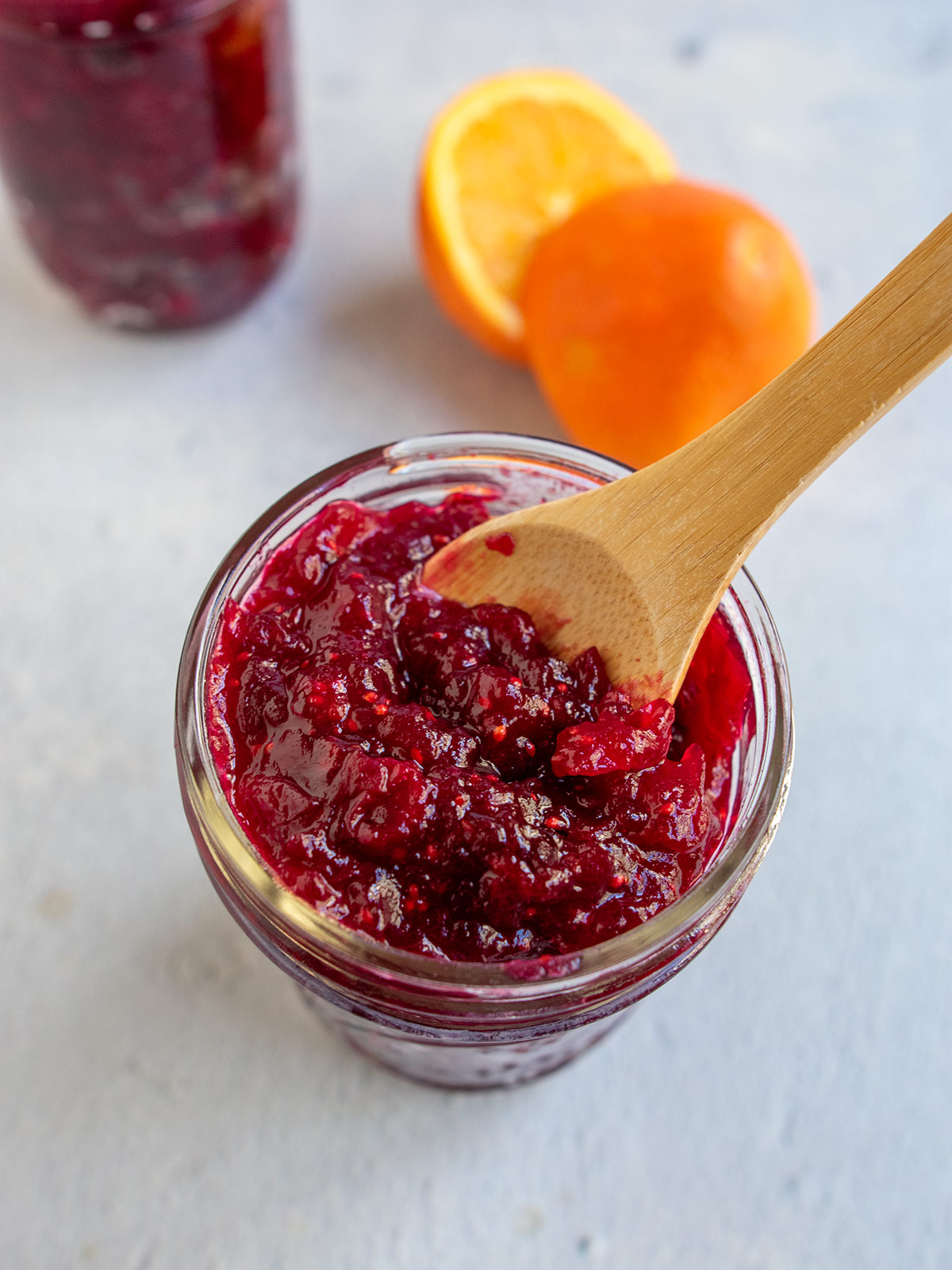 Jar of cranberry orange jam with a wooden spoon and a cut orange in the background on a light blue mat.