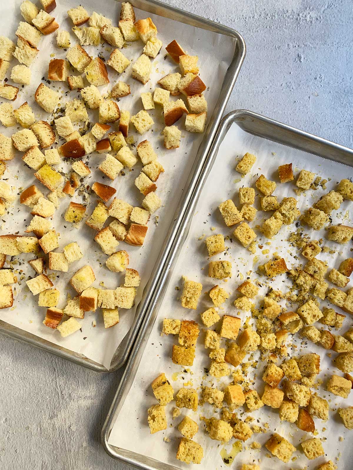 Two trays of challah and cornbread cubes with seasoning added shown at an angle.