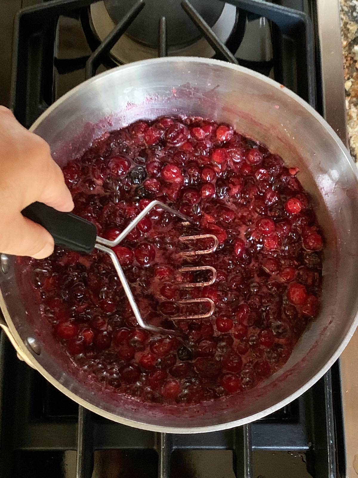 Hand holding potato masher mashing the cranberries in the pot.