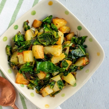 Greens and potatoes in a polka dot bowl with a copper spoon on a striped napkin.